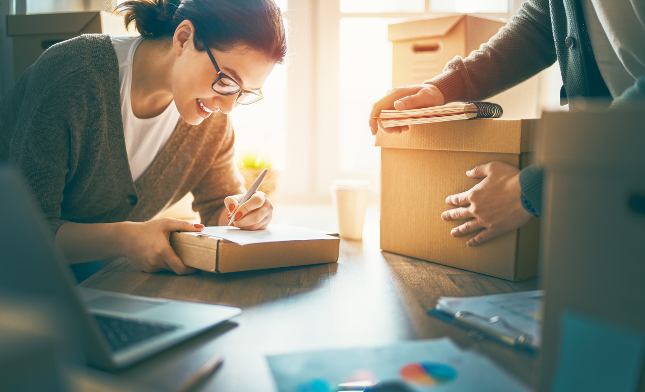 En kvinna skriver leende adressuppgifter på ett paket, medan en annan person håller i en kartong bredvid.