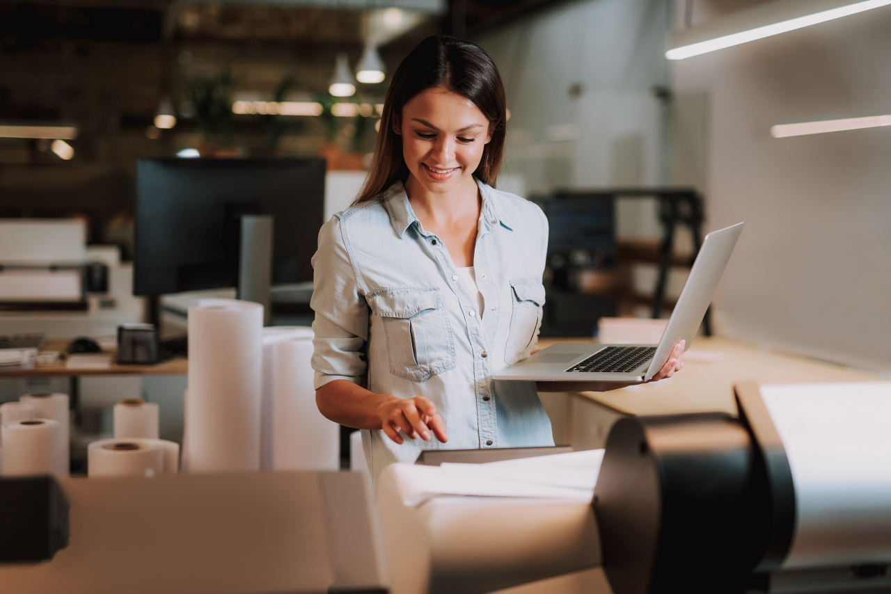 En kvinna står i en kontorsmiljö, använder en stor skrivare och håller en bärbar dator i den andra handen.