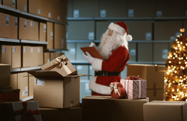 Jultomten granskar önskelistan i ett lager fullt av paket, med en julgran i bakgrunden. I förgrunden syns en öppen låda och julklappar.