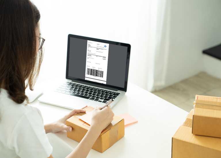 Kvinna skriver en anteckning på ett paket och använder Shipits fraktverktyg på sin bärbara dator.